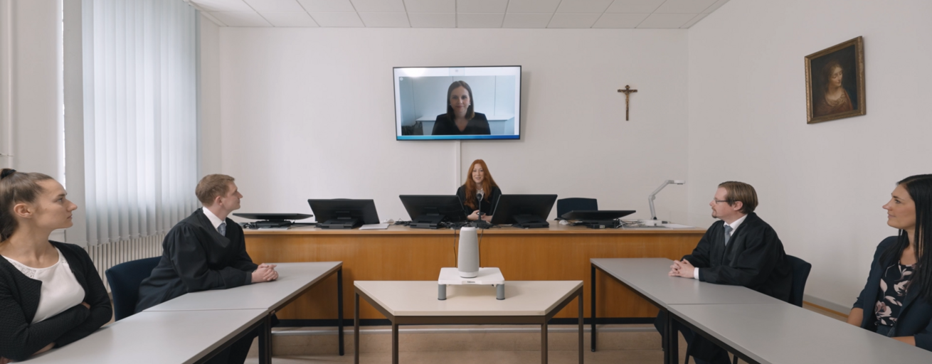 Screenshot aus Video: Gerichtssaal mit Richterin in der Mitte, zwei Parteien links und rechts und einer Frau zugeschaltet über Bildschirm