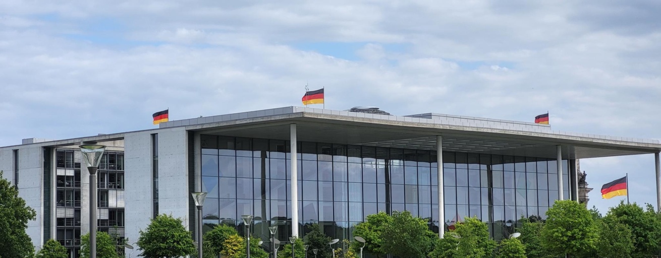 Man sieht ein Gebäude mit einer Glasfront vor einem blauen Himmel. Auf dem Dach wehen mehrere Deutschlandflaggen. Vor dem Gebäude laufen einige Menschen.