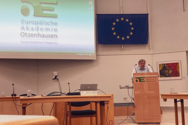 Prof. Weth steht am Rednerpult. Über ihm die Europaflagge, danaben auf der Leinwand das Logo der Europäischen Akademie Otzenhausen