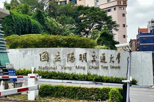 Man sieht eine Betonwand, auf der "National Yang Ming Chiao Tung University" in goldenen Buchstaben steht. Hinter der Wand befinden sich mehrere Bäume und einige mehrstöckige Häuser.