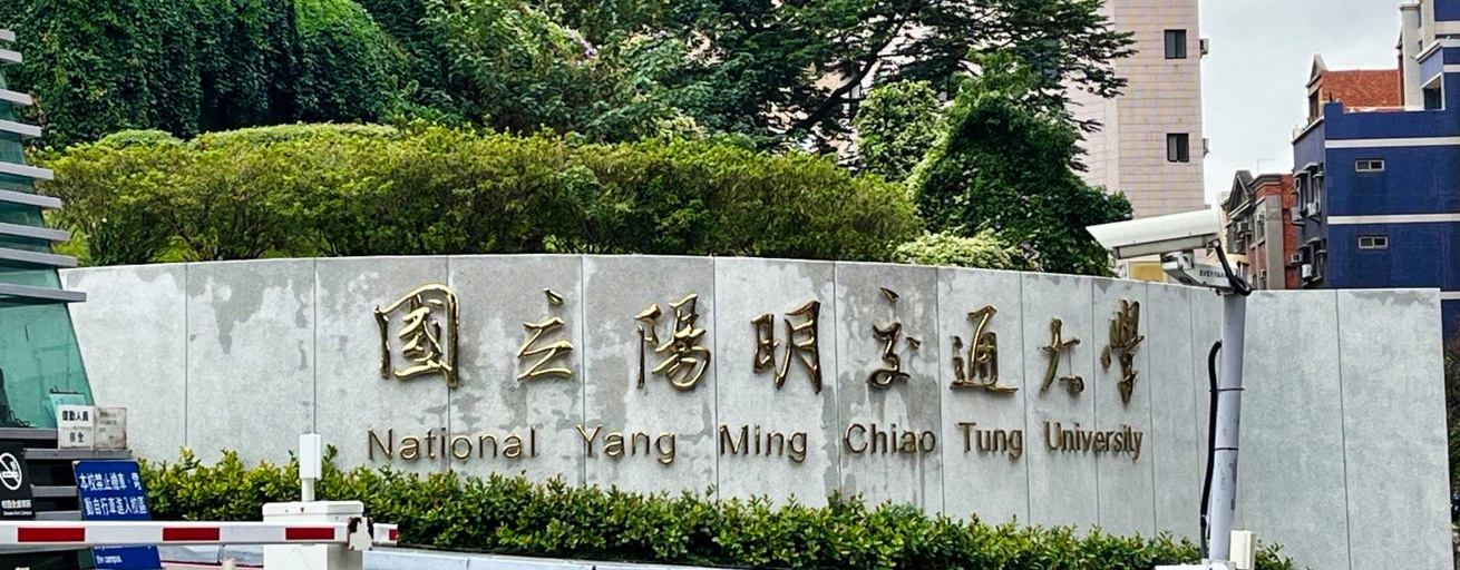 Man sieht eine Betonwand, auf der "National Yang Ming Chiao Tung University" in goldenen Buchstaben steht. Hinter der Wand befinden sich mehrere Bäume und einige mehrstöckige Häuser.