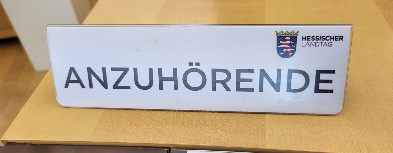 Auf einem Tisch aus hellem Holz steht ein Schild mit der Aufschrift "ANZUHÖRENDE". Oben rechts auf dem Schild sieht man das Logo des Hessischen Landtags.