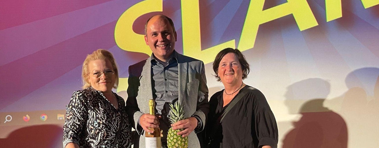 Drei Personen stehen vor einem bunten Hintergrund, auf dem mit gelber Schrift "Science Slam" geschrieben steht. Der Mann in der Mitte hält lächelnd eine Flasche und eine Ananas in den Händen.