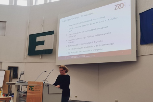 An einem Rednerpult steht eine Frau in einem schwarzen Kleid und hält einen Vortrag. Im Hintergrund sieht man eine PowerPoint-Präsentation mit dem Logo des ZRD.