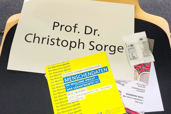 Man sieht ein Namensschild mit der Aufschrift "Prof. Dr. Christoph Sorge" neben einem gelben Flyer von der KI-Woche 2023 auf einem Stuhl liegen.