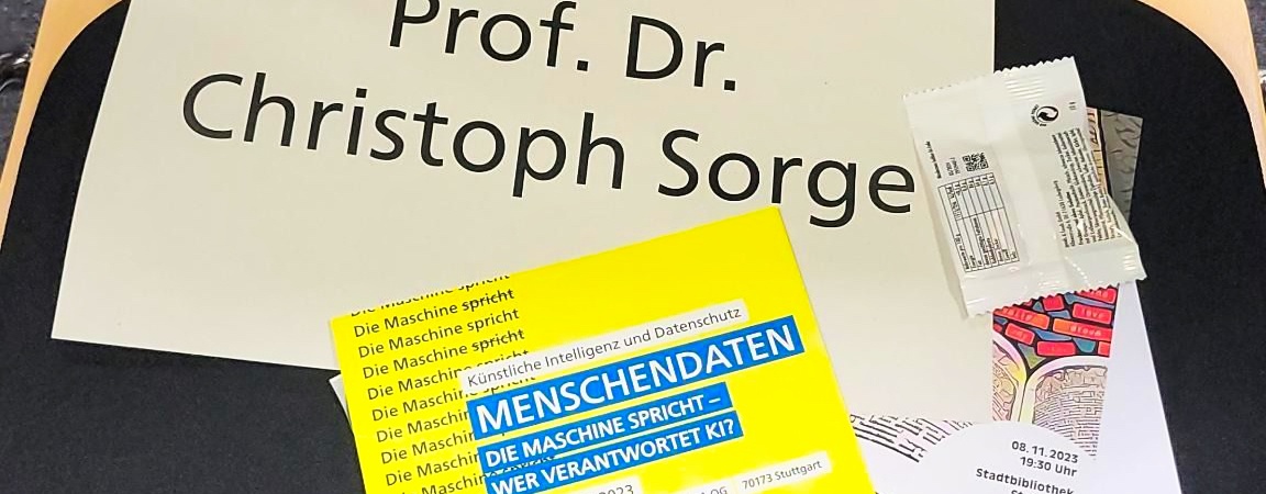 Man sieht ein Namensschild mit der Aufschrift "Prof. Dr. Christoph Sorge" neben einem gelben Flyer von der KI-Woche 2023 auf einem Stuhl liegen.