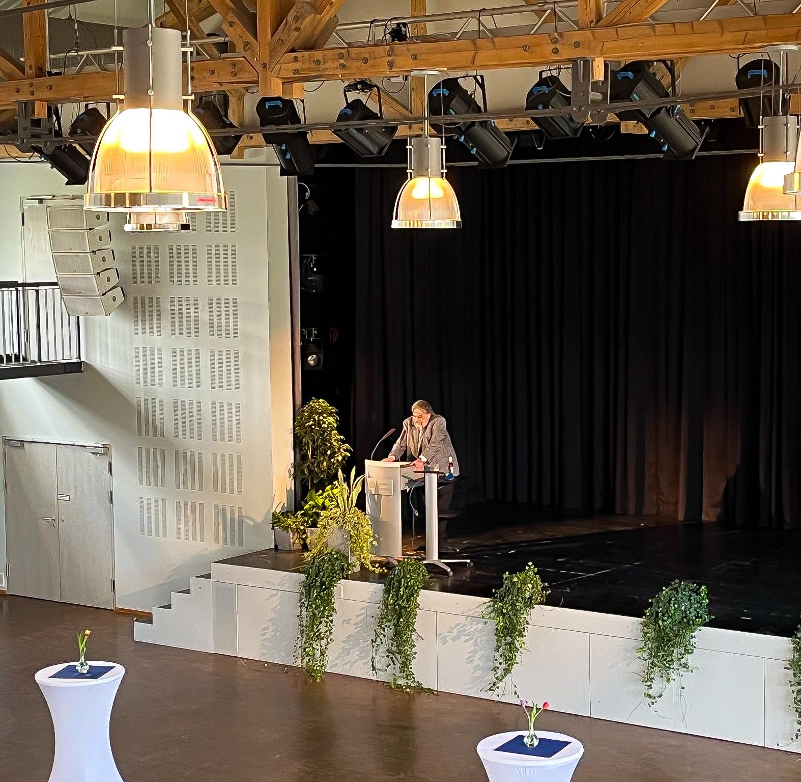 Prof. Stephan Weth spricht auf der Bühne der Aula der Universität des Saarlandes anlässlich der Feier zu seiner Emeritierung. Auf der Bühne stehen Grünpflanzen. Vor der Bühne stehen Stehtische.